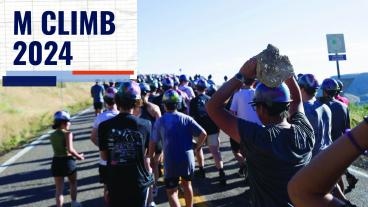 A large group of students, all wearing hard hats decorated with various colors, is hiking up a road during the M Climb tradition at Colorado School of Mines in 2024. The students are seen from the back, with one prominently holding a large rock above their head, which will be added to the famous hillside “M” on Mt. Zion. The image captures the early morning light and the sense of community and tradition as students participate in this historic event. The text "M CLIMB 2024" is overlaid on the left side.