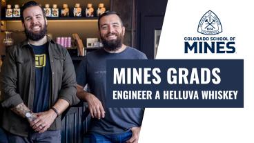 Two smiling Colorado School of Mines graduates stand side by side inside a modern distillery or bar. They both have beards and casual clothing—one wearing a gray T-shirt with a small logo, the other in a dark jacket over a graphic T-shirt. Shelves of whiskey bottles and other items are visible in the background. To the right, the Colorado School of Mines logo is displayed above bold text that reads "Mines Grads Engineer a Helluva Whiskey."