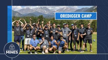 A group of first-year students at Colorado School of Mines pose outdoors during Ordigger Camp, with a backdrop of mountains and trees at YMCA of the Rockies. They are wearing Mines t-shirts and making playful gestures, showing excitement and team spirit. The banner on the top right reads "Oredigger Camp," and the Colorado School of Mines logo is placed in the lower left corner.