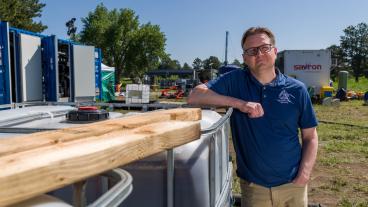 Chris Higgins stands in a field at Peterson Space Force Base in Colorado Springs, leaning on a large white tank with two wooden planks on top. In the background are large machines and testing equipment, related to PFAS research.