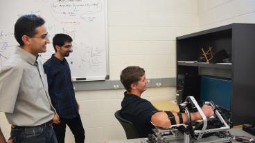 From left, mechanical engineering professor Ozkan Celik and graduate student Hossein Saadatzi watch as student David Long demonstrates the Wrist Gimbal.